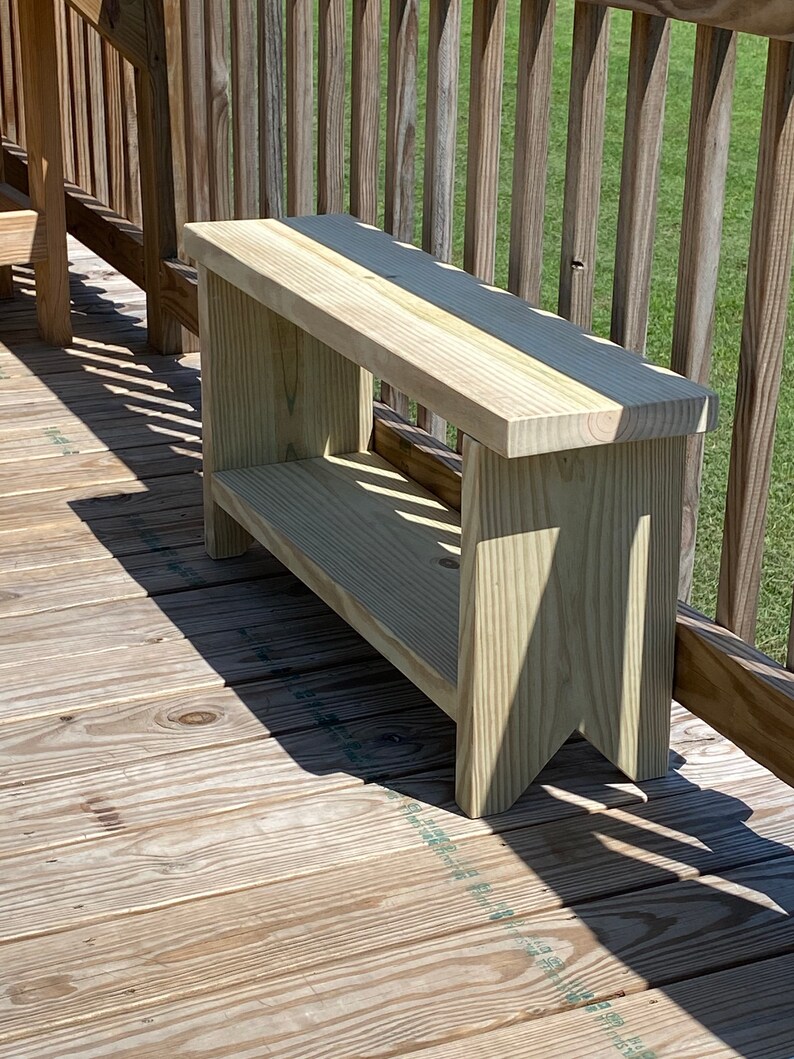 May include: A light-colored wooden bench with a rectangular seat and a lower shelf. The bench has a simple, sturdy design with angled legs. The wood grain is visible, and the bench is placed on a wooden deck.