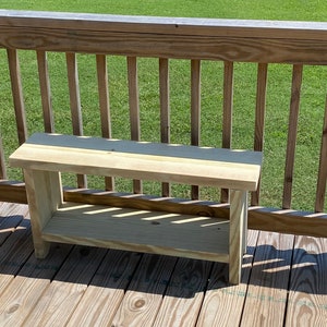 May include: A small, unfinished wooden bench with a shelf. The bench is made of light-colored wood and has a simple, rectangular design. It is sitting on a wooden deck with a wooden railing in the background.