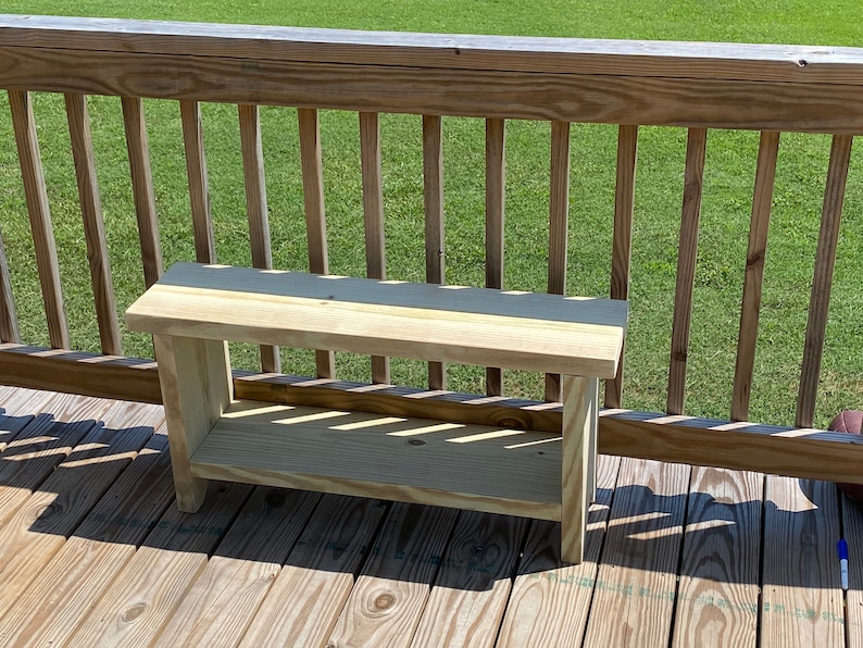 May include: A small, unfinished wooden bench with a shelf. The bench is made of light-colored wood and has a simple, rectangular design. It is sitting on a wooden deck with a wooden railing in the background.