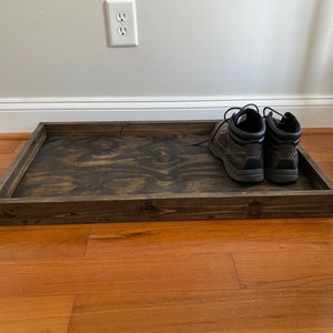 May include: A rectangular, dark stained wooden tray with raised edges. A pair of dark brown leather boots with laces are placed inside the tray. The tray sits on a hardwood floor.