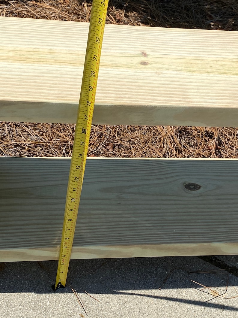 May include: A yellow measuring tape shows a wooden plank. The tape reads up to 24 inches. The wood is light-colored and appears to be new. The background is a mix of brown pine needles and a gray surface.