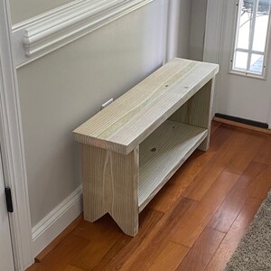 May include: A light-colored wooden bench with a lower shelf. The bench is rectangular with a simple design and is placed against a light gray wall. The bench is made of unfinished wood.