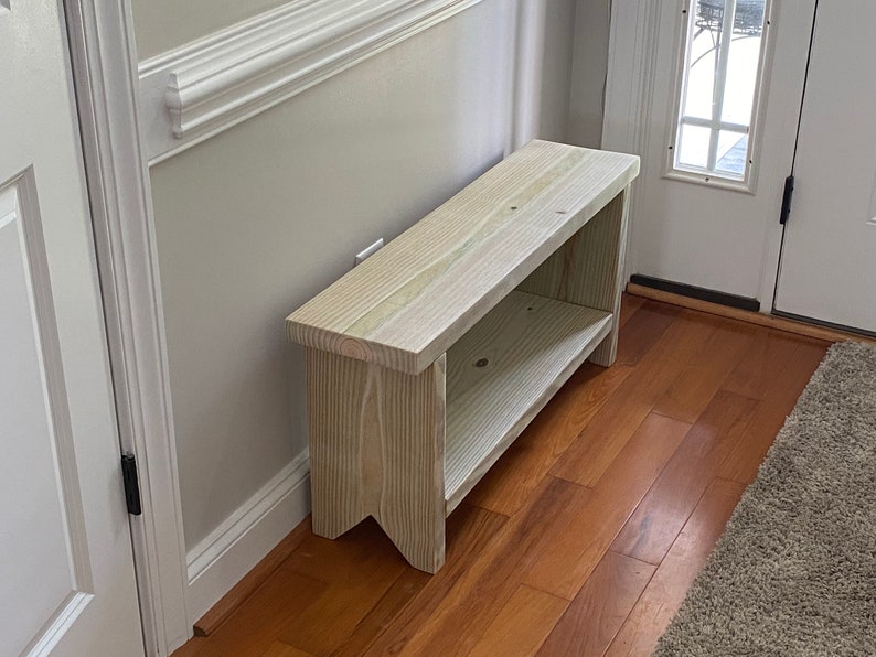 May include: A light-colored wooden bench with a lower shelf. The bench is rectangular with a simple design and is placed against a light gray wall. The bench is made of unfinished wood.