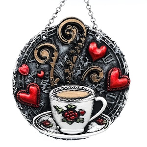 May include: A round, decorative wall hanging featuring a cup of coffee on a saucer, surrounded by red hearts. The background is a dark gray with gold accents and a clock-like design. A silver chain is attached for hanging.