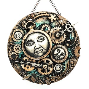 May include: A round, black, gold, and teal steampunk-style wall hanging with a clock face, gears, and two faces. The clock face has the number 12 on it.