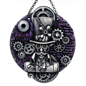May include: A silver and purple circular ornament with a steampunk design. The ornament features a man with a top hat and goggles, surrounded by gears and cogs. The ornament is hanging from a silver chain.