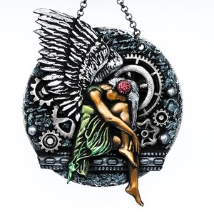 May include: A silver and black angel figurine with wings, wearing a green dress and a red flower in her hair. The angel is suspended from a chain and surrounded by gears and cogs.