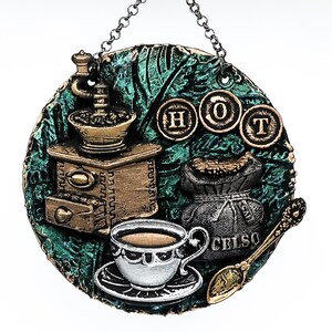 May include: A round, textured wall hanging with a coffee theme. It features a coffee grinder, a cup of coffee, a bag of coffee beans, and a spoon. The word "HOT" is displayed. The color scheme includes green, brown, gold, and silver.