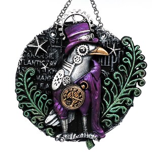 May include: A steampunk-style raven figurine with a purple top hat and coat, adorned with gears and a pocket watch. The raven is set against a black backdrop with text and star accents, framed by green foliage. A silver chain is attached for hanging.