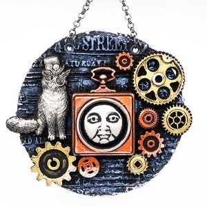 May include: A round, blue and orange, steampunk-style ornament with a cat wearing a top hat, gears, and a clock face. The ornament has the text "Old Street" and "Saturday" on it.