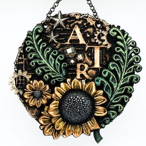 May include: A round, black and gold wall hanging with the word "ART" in gold lettering. The design includes sunflowers, gears, stars, and green leaf accents. A chain is attached for hanging.