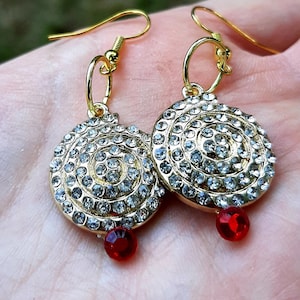 May include: A pair of gold earrings with a spiral design of clear rhinestones and a single red rhinestone dangling from each earring.