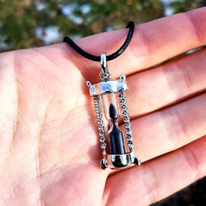 May include: A silver hourglass pendant necklace with a black cord. The hourglass is filled with black sand and has a silver chain around the top and bottom.