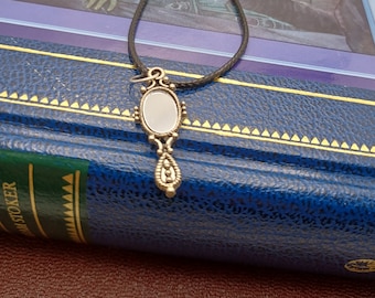 May include: A silver-toned pendant necklace with an oval-shaped, mirrored charm. The charm is suspended from a black cord and rests on a blue book. The pendant has ornate detailing and a small, teardrop-shaped element.