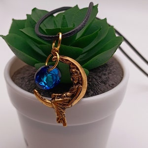 May include: A gold crescent moon pendant necklace with a blue crystal charm. The necklace is on a black cord and is displayed on a small potted plant.