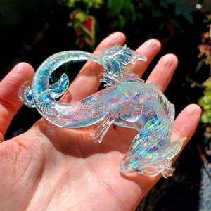 May include: A clear iridescent resin dragon figurine with glitter embedded in the resin. The dragon is curled up with its wings spread out.