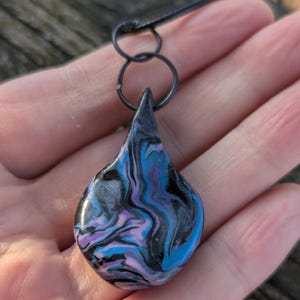 May include: A teardrop-shaped pendant with a swirling pattern of blue, purple, and black. The pendant is suspended from a black cord and attached to a black metal ring. The design is abstract and artistic.