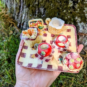 May include: A miniature ceramic tea set with a checkered picnic blanket design. The set includes a teapot, two cups, a basket of fruit, and a basket of strawberries.