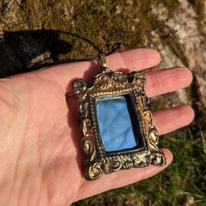 May include: A gold-toned ornate frame pendant necklace with a small mirror inside. The mirror reflects the sky and trees.