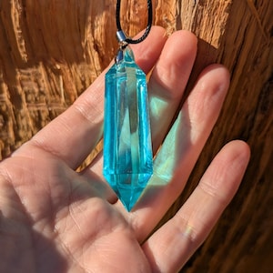 May include: A clear, blue crystal pendant with a pointed bottom and a silver-colored metal clasp. The pendant is suspended from a black cord necklace. The background is a textured, brown wooden surface.