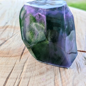 May include: A faceted, translucent resin sculpture with a mix of purple, green, and black hues. The object has a polished, crystalline appearance and sits on a wooden surface. The sculpture's interior features swirling color patterns.