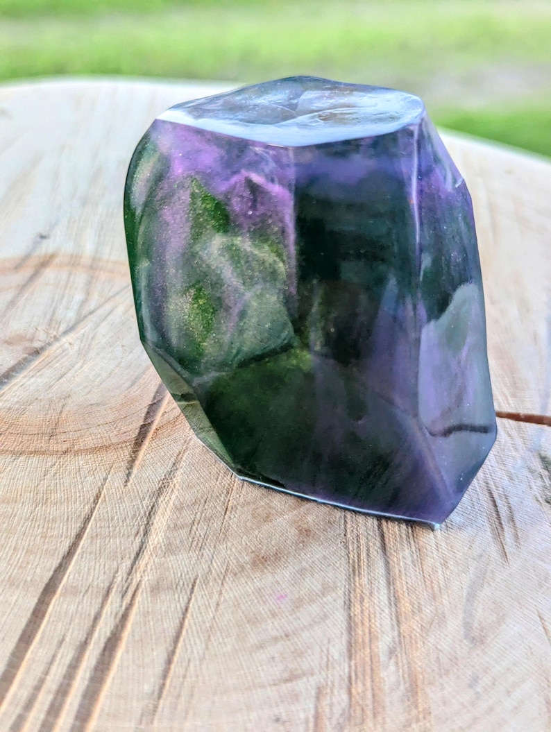 May include: A faceted, translucent resin sculpture with a mix of purple, green, and black hues. The object has a polished, crystalline appearance and sits on a wooden surface. The sculpture's interior features swirling color patterns.