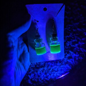May include: A pair of green glass bottle earrings with cork stoppers and silver hooks. The earrings are displayed on a white card with a hole in the top.