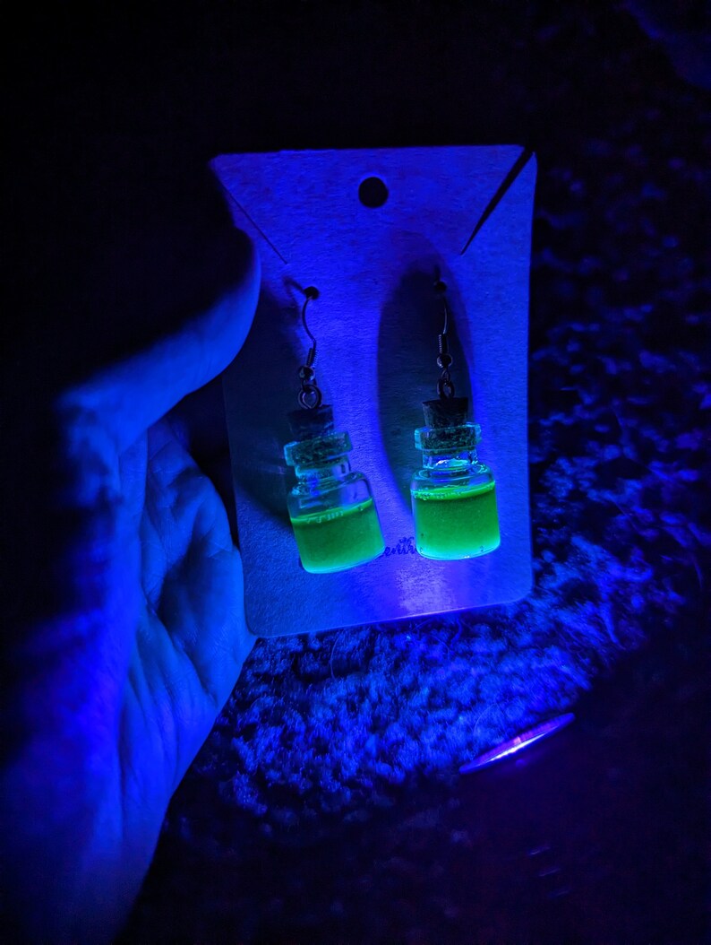 May include: A pair of green glass bottle earrings with cork stoppers and silver hooks. The earrings are displayed on a white card with a hole in the top.