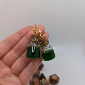May include: A pair of gold earrings with small glass bottles filled with green liquid and cork stoppers.