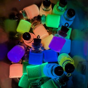 May include: A collection of small glass bottles filled with glowing, brightly colored sand. The bottles are arranged in a bowl and are illuminated by a blacklight.