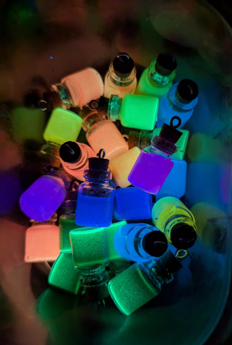 May include: A collection of small glass bottles filled with glowing, brightly colored sand. The bottles are arranged in a bowl and are illuminated by a blacklight.