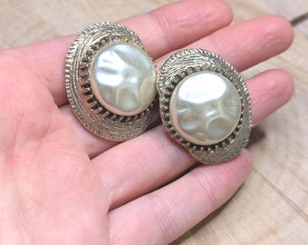 May include: A pair of vintage clip-on earrings. Each earring features a large, off-white, pearlescent center stone surrounded by a textured, gold-toned metal frame. The earrings are oval-shaped and have a classic, elegant design.