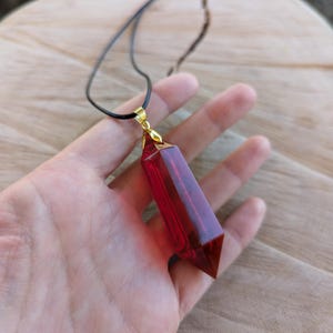 May include: A red crystal pendant on a black cord necklace. The pendant is shaped like a long, pointed teardrop.