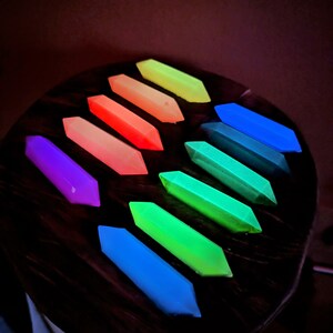 May include: A set of ten glow-in-the-dark, geometrically shaped, resin pieces in a rainbow of colors. The pieces are arranged in a semi-circle on a dark wooden surface.