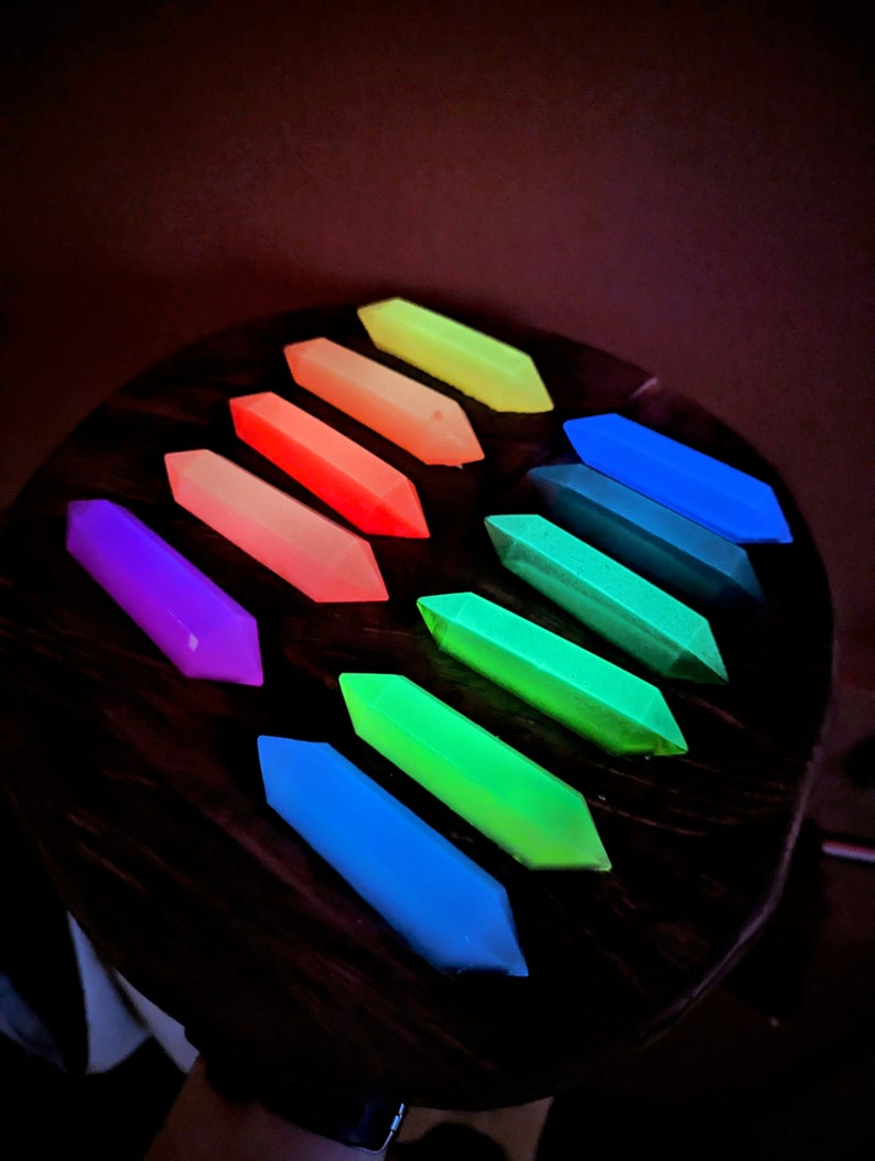 May include: A set of ten glow-in-the-dark, geometrically shaped, resin pieces in a rainbow of colors. The pieces are arranged in a semi-circle on a dark wooden surface.