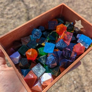 May include: A wooden box filled with colorful, iridescent dice. The dice are made of clear resin with embedded glitter and swirls of color. The box is filled with a variety of dice, including d4, d6, d8, d10, d12, and d20.