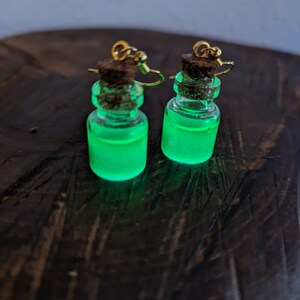 May include: A pair of green glow-in-the-dark bottle earrings with gold hooks and cork stoppers.