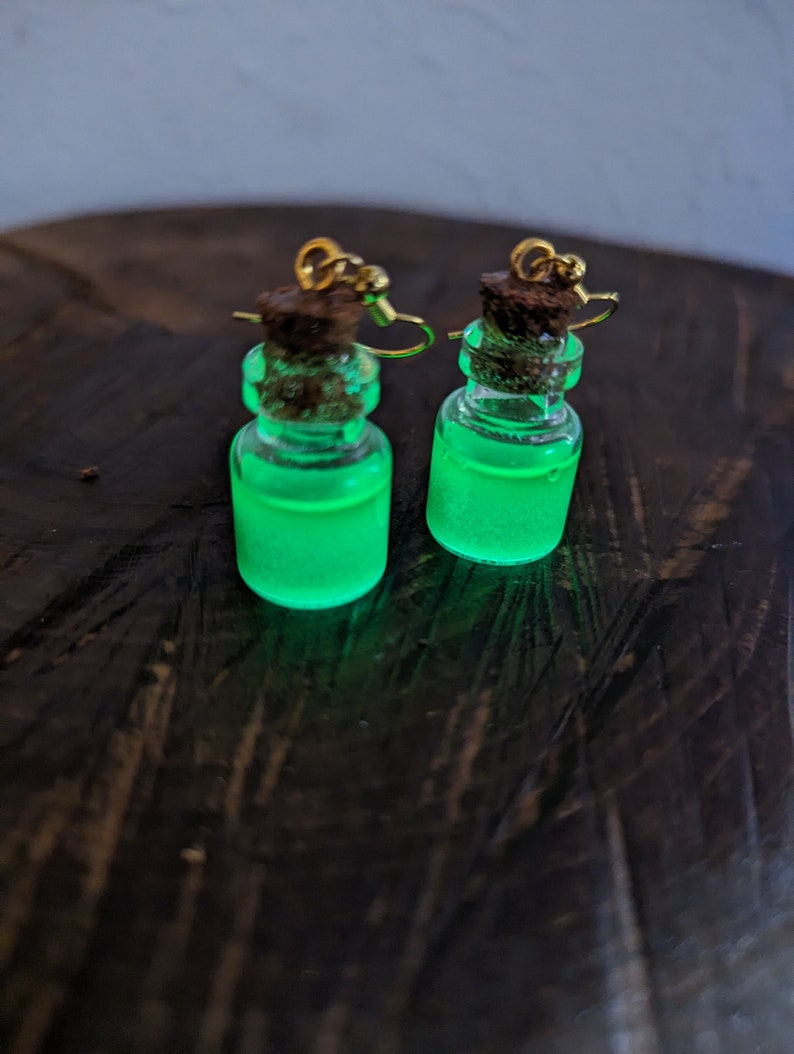 May include: A pair of green glow-in-the-dark bottle earrings with gold hooks and cork stoppers.