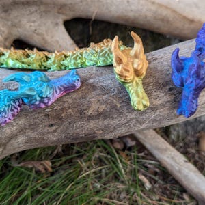 May include: A rainbow-colored dragon figurine made of plastic. The dragon is segmented and has a textured surface. The dragon is lying on a piece of driftwood.