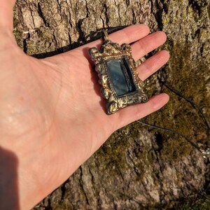 May include: A gold-toned ornate frame pendant necklace with a small mirror inside. The frame has a detailed design with floral motifs. The necklace is on a black cord.