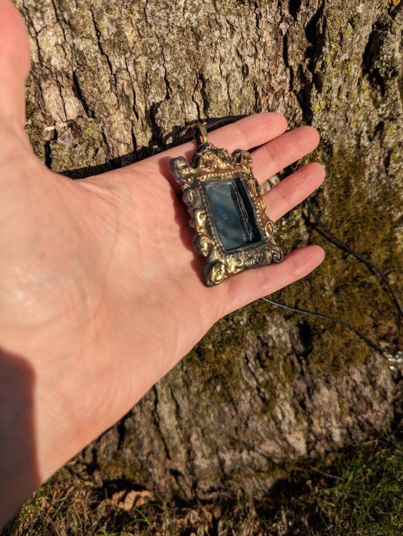 May include: A gold-toned ornate frame pendant necklace with a small mirror inside. The frame has a detailed design with floral motifs. The necklace is on a black cord.