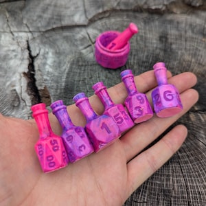 May include: A collection of miniature, hand-painted dice shaped like bottles, in shades of pink and purple. Each bottle features white numbers. A small mortar and pestle are also visible. The items are held in a hand.