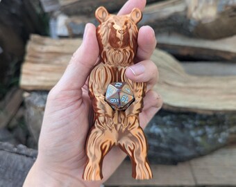 May include: A metallic copper-colored bear figurine standing on its hind legs, holding a multi-faceted polyhedral die between its front paws. The bear has detailed fur textures and a smooth, reflective finish, while the die features numbers.