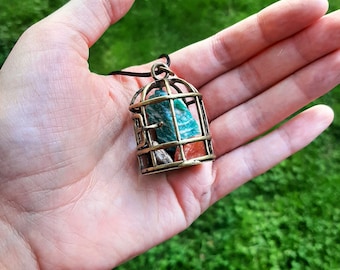 May include: A gold-toned birdcage pendant necklace with a green and brown stone inside. The necklace is on a black cord and is being held in a hand.