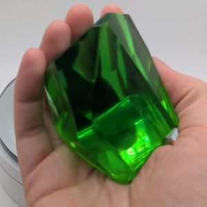 May include: A large, faceted, translucent green gemstone-shaped object with sharp, angular edges and a polished surface. The item displays deep emerald hues and light-reflecting planes, creating a clear, crystalline appearance.