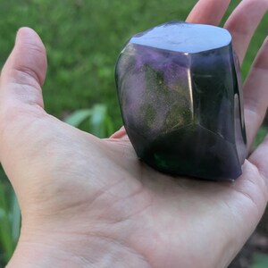 May include: A faceted, polished stone with a mix of purple, green, and black colors. The stone is held in an open hand, with a blurred green background.