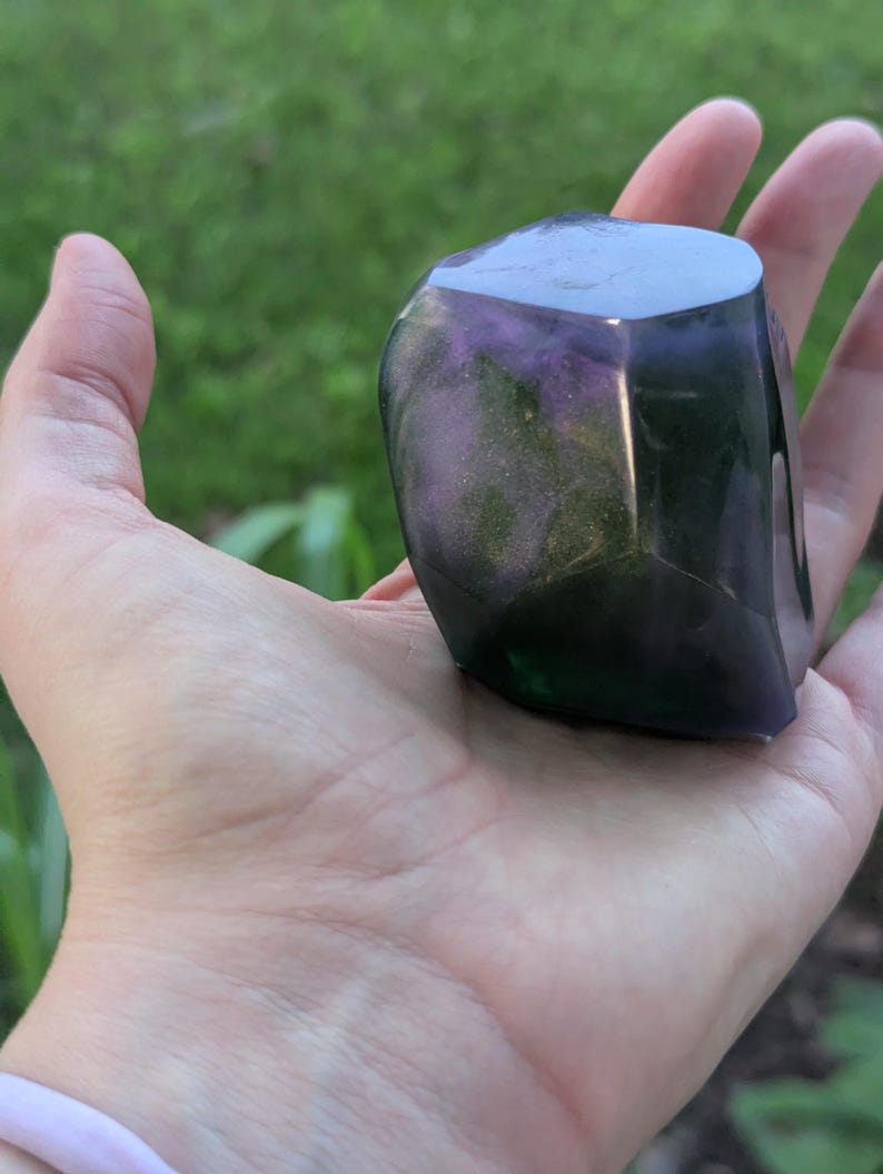 May include: A faceted, polished stone with a mix of purple, green, and black colors. The stone is held in an open hand, with a blurred green background.