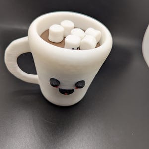 May include: A white ceramic mug filled with brown liquid and topped with white marshmallows. The mug has a cartoon face with black eyes and a red smile. The mug has a handle.