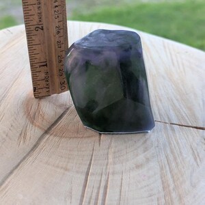 May include: A dark, faceted stone with purple and green hues, approximately 2.5 inches tall, is shown next to a wooden ruler. The stone has a polished appearance and sits on a light-colored wooden surface.