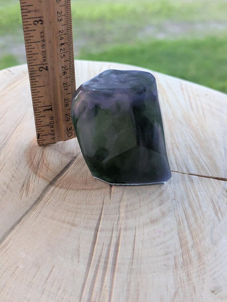 May include: A dark, faceted stone with purple and green hues, approximately 2.5 inches tall, is shown next to a wooden ruler. The stone has a polished appearance and sits on a light-colored wooden surface.